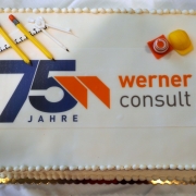Zweistöckige, weiße Torte mit 75 Jahre WERNER CONSULT Logo bedruckt. einem Baustellenhütchen und Sicherheitshelm rechts oben. Links oben sind ein Zirkel, Bleistifte und ein Lineal.