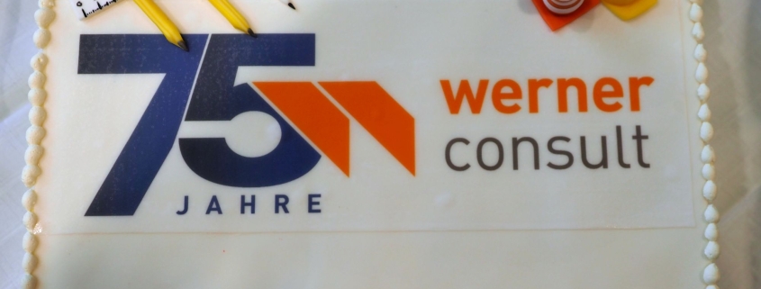 Zweistöckige, weiße Torte mit 75 Jahre WERNER CONSULT Logo bedruckt. einem Baustellenhütchen und Sicherheitshelm rechts oben. Links oben sind ein Zirkel, Bleistifte und ein Lineal.